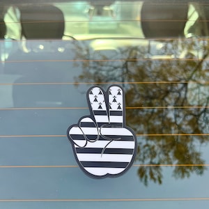 Peut inclure: Un autocollant en forme de signe de paix noir et blanc avec un design de drapeau breton. L'autocollant est sur une vitre de voiture.