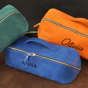 May include: Three suede-like travel bags in teal, blue, and orange. Each bag has a top handle, a gold zipper, and a name or initial. The teal bag has a "D" initial with a laurel wreath. The blue bag says "ANNA". The orange bag says "Olivia".