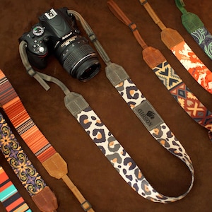 Può includere: Una fotocamera Nikon nera con un obiettivo è mostrata con diversi cinturini per fotocamera a fantasia. I cinturini presentano disegni come stampa leopardata, motivi geometrici e motivi floreali. I cinturini sono in vari colori, tra cui arancione, marrone e verde.
