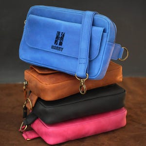 May include: A stack of four leather crossbody bags in blue, brown, black, and pink. The blue bag on top has the word "HARRY" printed on it. Each bag has a strap and gold-colored hardware.