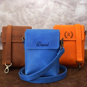 Puede incluir: Tres bolsos cruzados de cuero en marrón, azul y naranja. El bolso marrón tiene grabado "OLIVIA". El bolso azul tiene "David" en cursiva. El bolso naranja tiene un monograma. Cada bolso tiene una correa para el hombro y herrajes dorados.