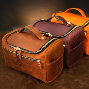 May include: Three leather travel bags in shades of brown, burgundy, and orange. Each bag has a top handle, a zippered closure with a black zip, and a small front pocket. The front bag has an initial and decorative design.