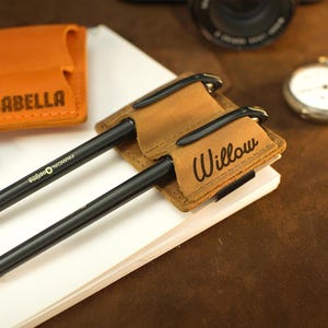 May include: A close-up of personalized leather pen holders with black pens. One holder is tan with the name "Willow", another is orange with "Isabella", and a blue one with "Amelia". A pocket watch and camera are in the background.
