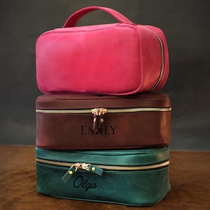May include: Three leather cosmetic bags stacked on top of each other. The top bag is a bright pink, the middle bag is brown with "EMILY" embossed, and the bottom bag is teal with "Olga" embossed. Each bag has a gold zipper and a handle.
