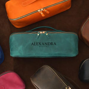 May include: A collection of leather travel cases in various colors, including teal, orange, brown, blue, black, and pink. The teal case in the center has the text "ALEXANDRA MOORE". Each case features a zipper closure and a handle.