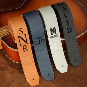 Può includere: Quattro cinturini per chitarra personalizzati in vari colori: marrone, blu, bianco e grigio. Ogni cinturino presenta testo personalizzato in nero, inclusi iniziali e nomi. I cinturini sono esposti contro una chitarra.