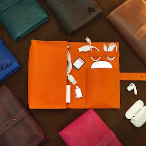Op de afbeelding: Een open oranje leren tech-organizer met witte kabels, een muis en een geheugenkaart. Gesloten leren organizers in verschillende kleuren, waaronder blauw, groen en bruin, zijn rond de open organizer gerangschikt. Elke gesloten organizer heeft een gegraveerde naam.
