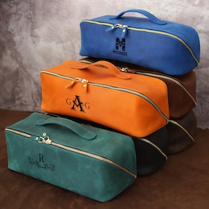 May include: A stack of rectangular leather toiletry bags in various colors: teal, orange, brown, and blue. Each bag has a gold zipper, a handle, and personalized initials or a name. The bags are approximately 10 inches long.