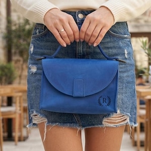 May include: A vibrant blue handbag with a top handle and a flap closure. The bag features a subtle embossed design on the front. The person is wearing a denim skirt. The background includes outdoor seating and potted plants.