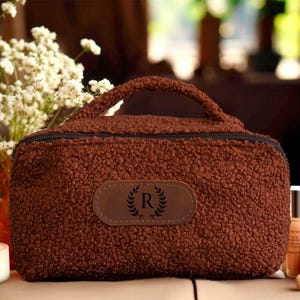 May include: A brown, textured cosmetic bag with a handle and a zip closure. The bag has a leather patch with a laurel wreath and the letter "R". Beauty products and a vase of white flowers are in the background.