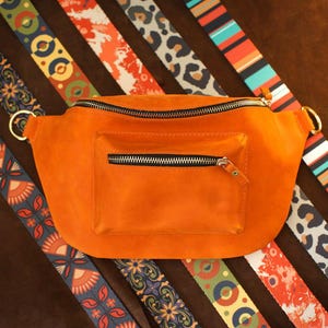 Könnte beinhalten: Orangefarbene Bauchtasche aus Wildleder mit einer Reißverschlusstasche vorne und einem Reißverschluss oben. Die Tasche hat goldfarbene Ringe zum Anbringen eines Riemens. Mehrere gemusterte Riemen in verschiedenen Farben und Designs sind um die Tasche angeordnet.