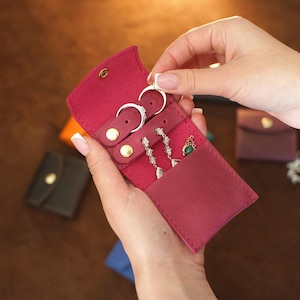 May include: A burgundy leather jewellery travel case with gold snap closures. The case is open, displaying rings and earrings. Other small jewellery cases are visible in the background.
