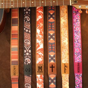 May include: Acoustic guitar with a selection of colorful guitar straps. Straps feature various patterns, including stripes, paisley, and floral designs. Some straps have personalized names like "Jacob," "Michael," "Maria," and "Patricia."