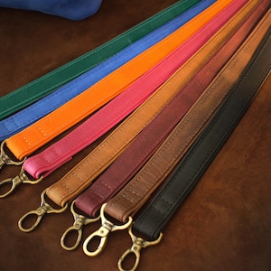 May include: A collection of colorful leather straps with brass-colored clasps. The straps are in various colors, including green, blue, orange, pink, brown, and black. The straps are approximately 1 inch wide and are designed for use with bags or other accessories.