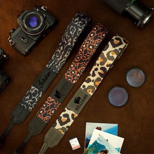 May include: Three camera straps with leopard print patterns in black, brown, and tan, each with a black leather end. The straps are personalized with names: "Jordan", "Martin", and "Williams". Two cameras and lenses are visible.