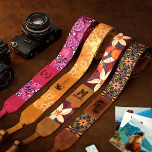 Pode incluir: Quatro correias de câmera estampadas com desenhos florais em rosa, laranja e roxo, ao lado de duas câmeras pretas. As correias têm etiquetas personalizadas com nomes e iniciais. Algumas fotos impressas são visíveis.
