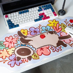 Peut inclure: Un tapis de souris blanc avec un chat de dessin animé dormant dans un lit de fleurs colorées. Le chat est brun et blanc avec un nez rose. Les fleurs sont roses, violettes, jaunes et vertes. Il y a une souris d'ordinateur blanche sur le tapis de souris.