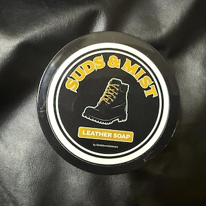 Könnte beinhalten: Runder schwarzer Behälter mit Lederseife. Die Worte "SUDS & MIST" sind gelb. Ein schwarzer Stiefel mit gelben Schnürsenkeln befindet sich in der Mitte. Darunter steht "LEATHER SOAP".