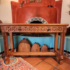 May include: A light brown wooden console table with two drawers and carved floral designs. The table has a lower shelf and is supported by four legs. Three wooden decorative objects are beneath the table. A colorful rug is on the floor.