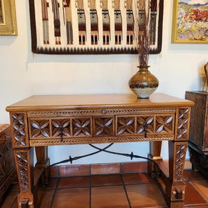 May include: A wooden console table with carved floral designs on the drawer fronts and legs. The table has a light brown finish and a decorative metal support. A ceramic vase with dried plants sits on top of the table.
