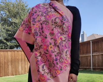 Op de afbeelding: Een roze sjaal met een bloemenpatroon in roze, gele en groene tinten. De sjaal heeft een lichtroze rand en kwastjes. De sjaal is gedrapeerd over een zwarte outfit.