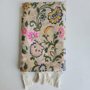 May include: A beige scarf with a floral pattern in pink, yellow, and blue. The scarf has a fringe at the bottom. The floral design includes leaves and stems in green and dark green.