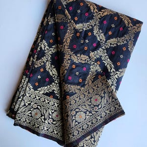 May include: A black fabric textile featuring an intricate gold metallic woven pattern of floral vines and leaves. The design includes small scattered square motifs in vibrant orange and pink, finished with a wide, detailed decorative border.