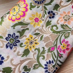 May include: Close-up of a white fabric with a detailed floral pattern. The design features large flowers in shades of yellow, pink, and blue, with green leaves and gold accents. The fabric is folded, revealing a fringe.