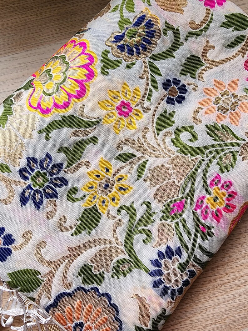 May include: Close-up of a white fabric with a detailed floral pattern. The design features large flowers in shades of yellow, pink, and blue, with green leaves and gold accents. The fabric is folded, revealing a fringe.