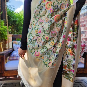 May include: A cream-colored shawl with a floral pattern in pink, yellow, green, and blue. The shawl has a gold border and white tassels. The floral design is intricate and covers the entire shawl.