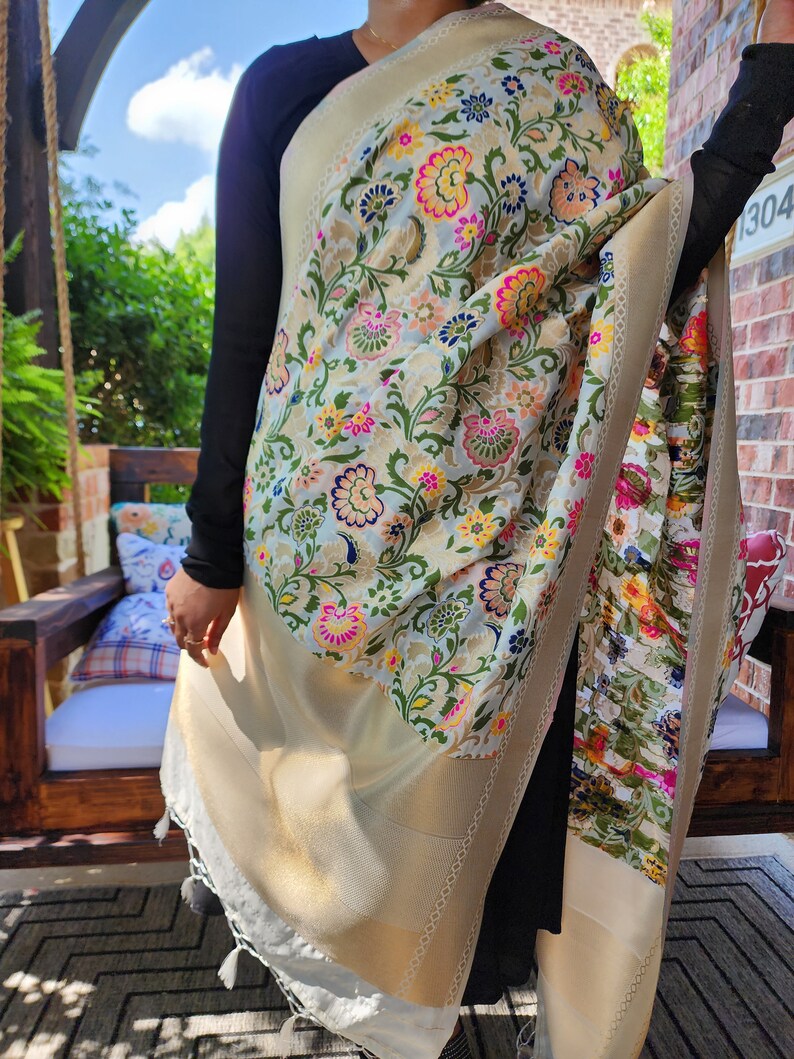 May include: A cream-colored shawl with a floral pattern in pink, yellow, green, and blue. The shawl has a gold border and white tassels. The floral design is intricate and covers the entire shawl.