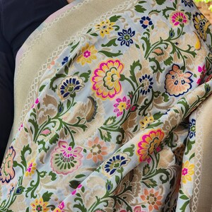 May include: A light-colored textile with a floral pattern in pink, yellow, blue, and orange. The fabric has a gold border with a geometric design. The textile is draped over a dark-colored garment.
