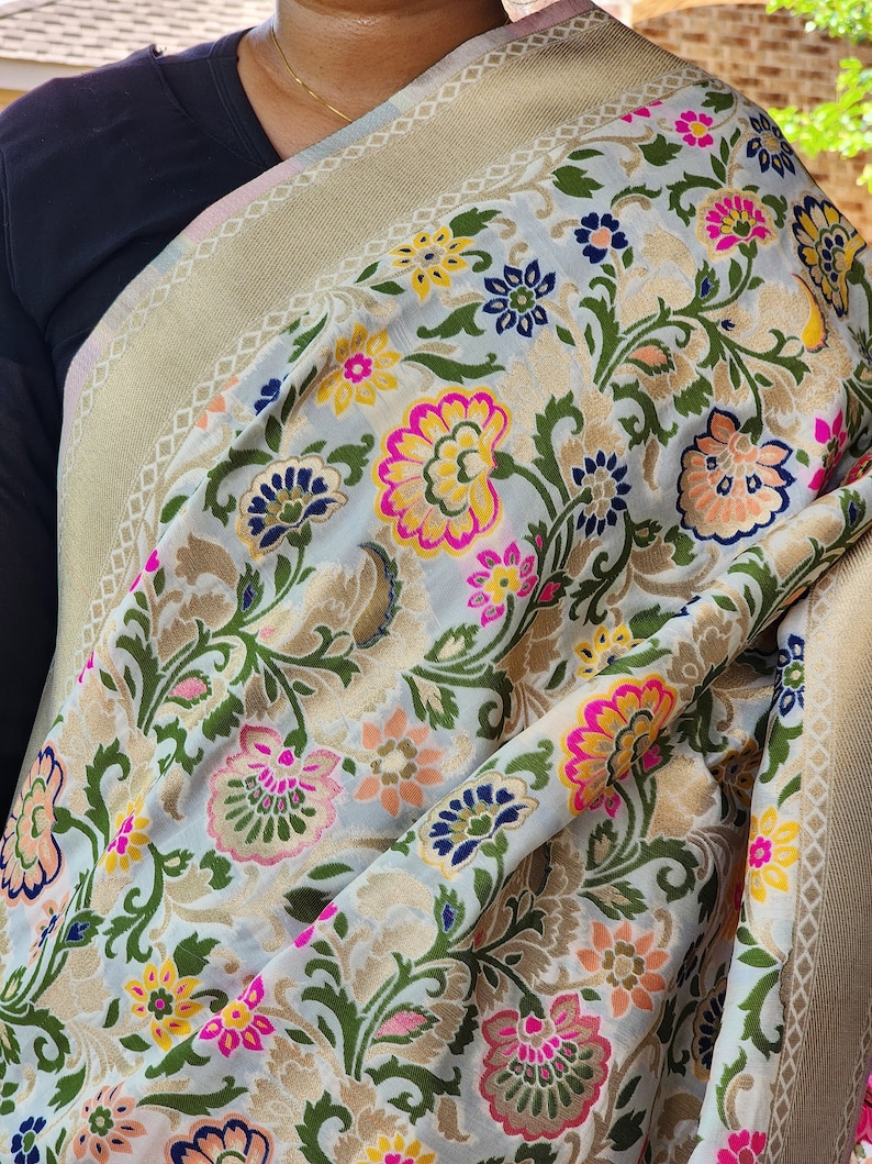 May include: A light-colored textile with a floral pattern in pink, yellow, blue, and orange. The fabric has a gold border with a geometric design. The textile is draped over a dark-colored garment.