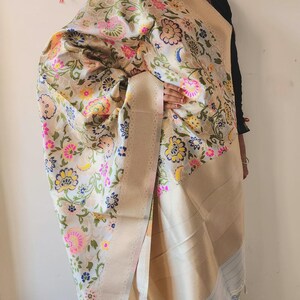 May include: A cream-colored shawl with a floral pattern in pink, yellow, and blue. The shawl has a beige border and is draped over the shoulder. The floral design is intricate and detailed, creating a visually appealing textile.