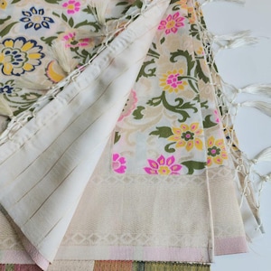 May include: Cream-colored textile with a floral pattern in pink, yellow, and green. The fabric has a striped section and is trimmed with tassels. The design features a repeating floral motif, creating a decorative and elegant look.