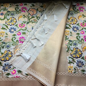May include: A folded textile with a floral pattern in gold, pink, yellow, and blue on a light blue background. A section of the textile is folded over, revealing a beige fabric with a geometric border and white tassels.