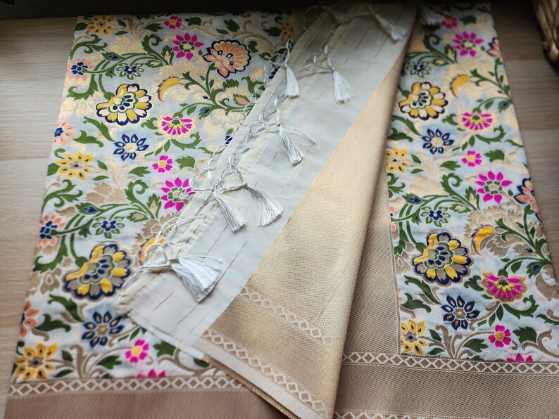 May include: A folded textile with a floral pattern in gold, pink, yellow, and blue on a light blue background. A section of the textile is folded over, revealing a beige fabric with a geometric border and white tassels.