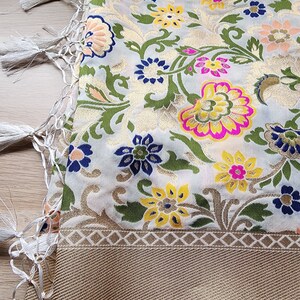 May include: A detailed shot of a scarf with a floral pattern. The scarf features a cream-colored background with gold, pink, yellow, blue, and green floral designs. The border is a gold color with a geometric pattern, and the scarf has white tassels.
