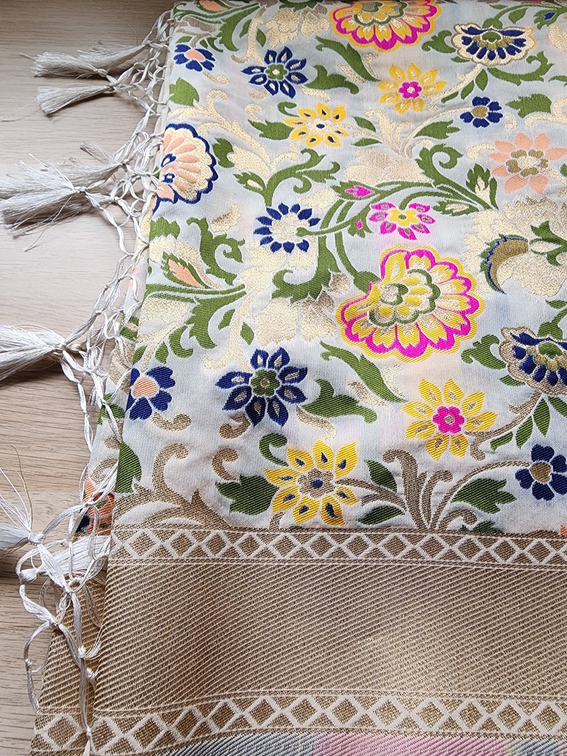 May include: A detailed shot of a scarf with a floral pattern. The scarf features a cream-colored background with gold, pink, yellow, blue, and green floral designs. The border is a gold color with a geometric pattern, and the scarf has white tassels.