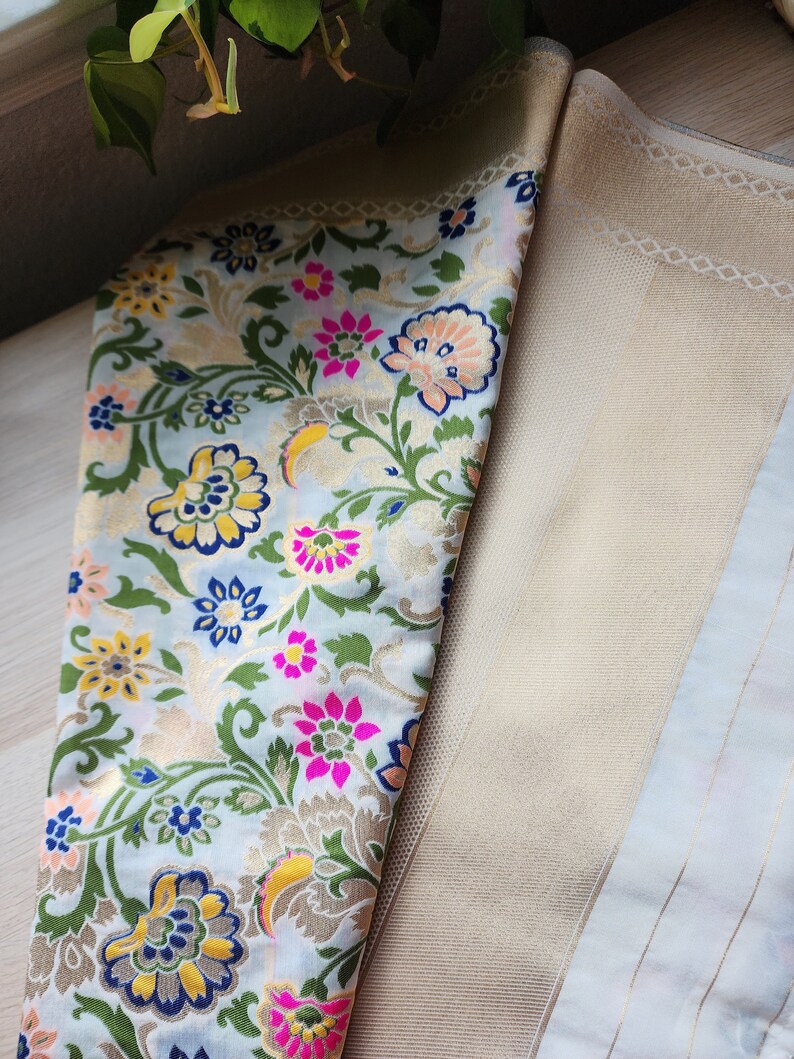 May include: Close-up of a folded fabric with a floral pattern. The fabric features a light blue background with colorful flowers in pink, yellow, and blue, and green leaves. The fabric on the right is a solid beige color with a subtle pattern.