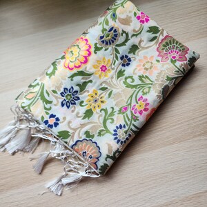 May include: Folded cream-colored scarf with a floral pattern in pink, yellow, blue, and green. The scarf has a gold metallic sheen and white tassels along one edge. The fabric appears to be a woven material.
