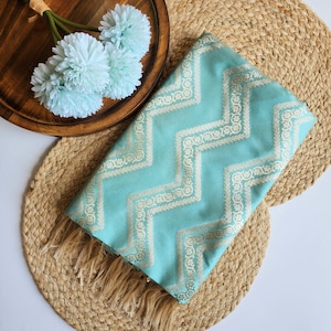 May include: A folded light turquoise fabric featuring a repeating cream-colored chevron pattern with intricate floral details. The textile has a smooth finish and includes fringe detailing along the bottom edge, showcasing a consistent geometric design.
