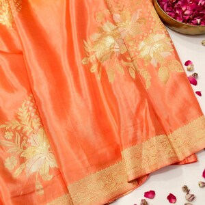 May include: A vibrant orange fabric featuring intricate gold floral patterns woven into the material. The textile displays a wide, ornate gold border along the bottom edge with detailed geometric and botanical motifs, creating a rich, textured appearance.