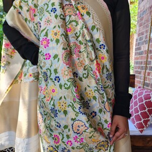 May include: A floral patterned shawl with a light blue background, featuring pink, yellow, and blue flowers. The shawl has a gold border and white tassels. The fabric is draped over the shoulders.