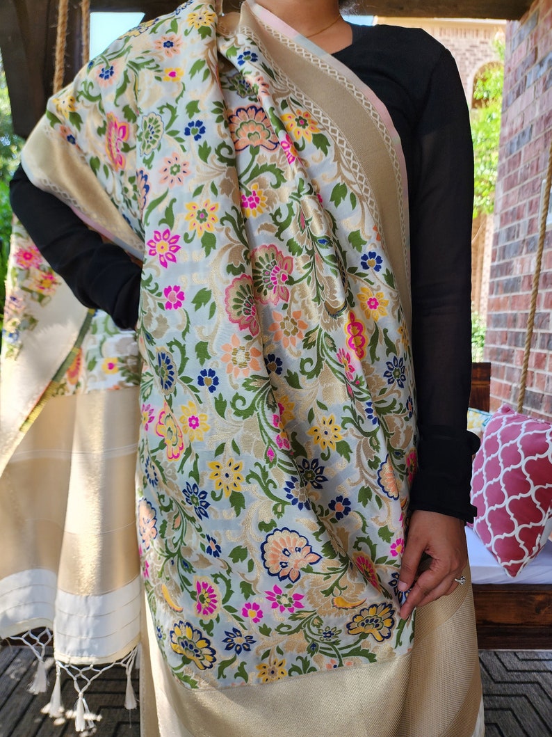May include: A floral patterned shawl with a light blue background, featuring pink, yellow, and blue flowers. The shawl has a gold border and white tassels. The fabric is draped over the shoulders.