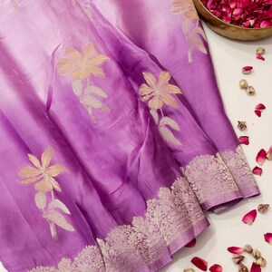 May include: A lavender and purple ombre saree featuring a wide, intricate silver metallic border with floral patterns and scattered gold-toned embroidered flower motifs across the fabric, accompanied by a brass bowl filled with pink rose petals.