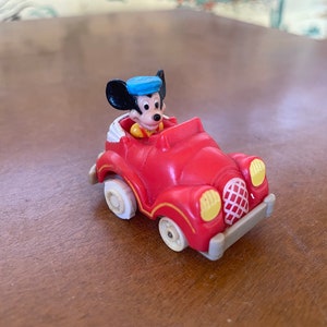 Puede incluir: Un coche de juguete rojo con una figura de Mickey Mouse al volante. Mickey lleva una camisa amarilla y un sombrero azul. El coche tiene faros delanteros amarillos y una parrilla blanca.