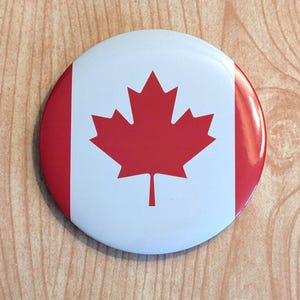 May include: A round button with a white background and a red maple leaf in the centre. The button has a red border.