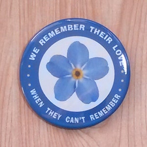 Può includere: Bottone blu e bianco con un fiore di nontiscordardime e il testo "Ricordiamo il loro amore quando loro non possono ricordare."