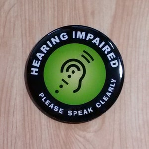 May include: A black and green round button with the text "Hearing Impaired Please Speak Clearly" and a stylised image of an ear with sound waves.